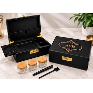 May include: Two black wooden storage boxes with gold-coloured combination locks. One box is open, revealing internal compartments and a sliding tray. The closed box features the name ASH in gold lettering. Includes three glass jars with bamboo lids.