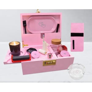 Puede incluir: Una caja de madera rosa con varios accesorios, incluyendo una bandeja con diseños de corazones, una vela y un molinillo. La caja tiene una cerradura de combinación y un cierre dorado. También se ve un cenicero con diseños de corazones.