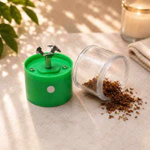 May include: A portable electric herb grinder with a bright green cylindrical base, a silver metal blade assembly on top, and a clear plastic container lying on its side next to it, containing a small pile of finely shredded brown dried herbs.