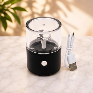 May include: A portable electric food chopper with a matte black cylindrical base, a clear plastic container, and a silver metal blade assembly. A white circular power button is on the front. A white USB charging cable is included with the device.