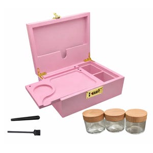 May include: A pink wooden box with a hinged lid, gold-colored hardware, and a combination lock. It includes a built-in tray with a circular cutout and two small compartments. Three glass jars with wooden lids and two black tools are also included.