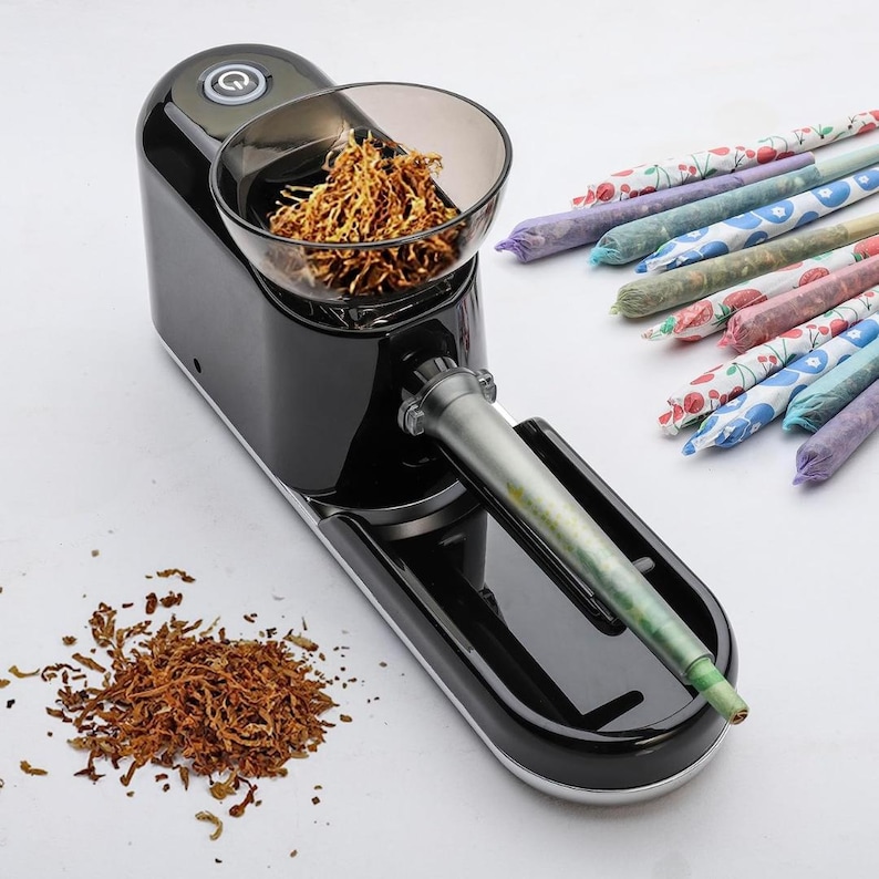May include: Black electric rolling machine with a clear bowl filled with shredded tobacco. A tray holds the rolling tube. Several pre-rolled joints with colorful patterned paper are arranged to the right.