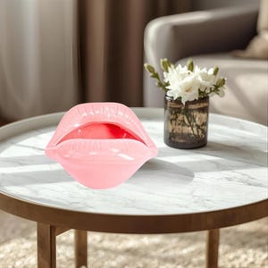May include: A glossy, light pink ceramic sculpture shaped like a pair of open lips. The object features a smooth, reflective surface with defined contours representing the upper and lower lips, creating a hollow opening in the center of the form.