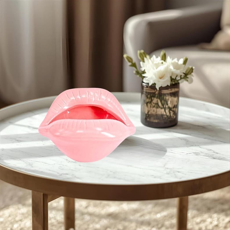 May include: A glossy, light pink ceramic sculpture shaped like a pair of open lips. The object features a smooth, reflective surface with defined contours representing the upper and lower lips, creating a hollow opening in the center of the form.