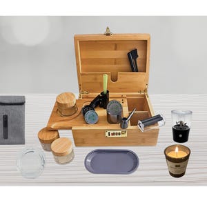 Peut inclure: Un coffret en bois avec divers accessoires, dont un broyeur, des bocaux, un plateau et une bougie. La boîte est ouverte, révélant des compartiments de rangement. Une pochette grise et un petit mixeur sont également présents.