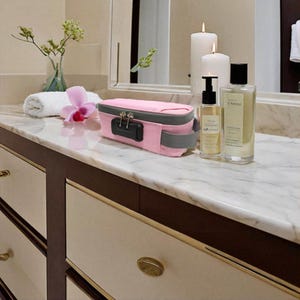May include: A rectangular pink storage pouch with a grey zipper and a black combination lock mechanism on the front. The pouch features a grey side handle and is constructed from a textured fabric material, designed for secure storage of small items.