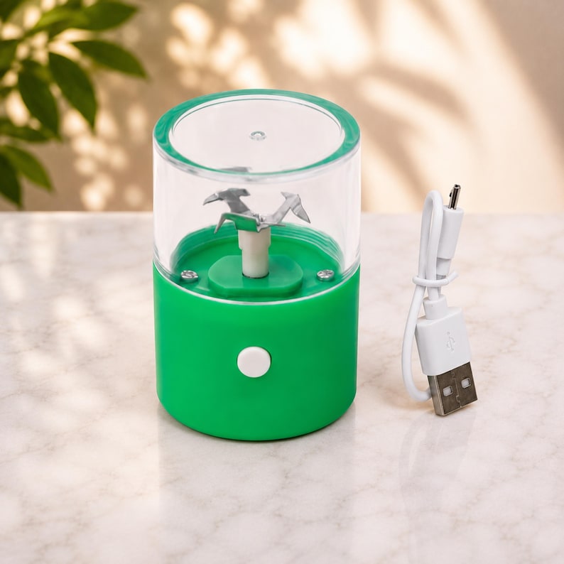 May include: A portable green electric food chopper with a clear plastic container and a metal blade assembly. The device features a white power button on the front and includes a white USB charging cable with a standard connector for power supply.