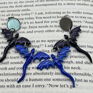 May include: A pair of unique earrings featuring a bat design. The earrings have a dark blue and black color scheme, with a shiny finish. The earrings are set against a backdrop of text from a book.