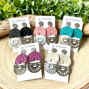 May include: A selection of handmade earrings in various colours, presented on white cards. Each pair has a textured, semi-circular top in black, pink, teal, purple, and cream, with a silver, sunburst-shaped bottom. The brand name "Blue Holly Boutique" is printed on each card.