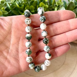 May include: A beaded bracelet featuring white and green moss agate stones. The round beads are interspersed with small gold-colored spacers. The bracelet is displayed against a background of green foliage.