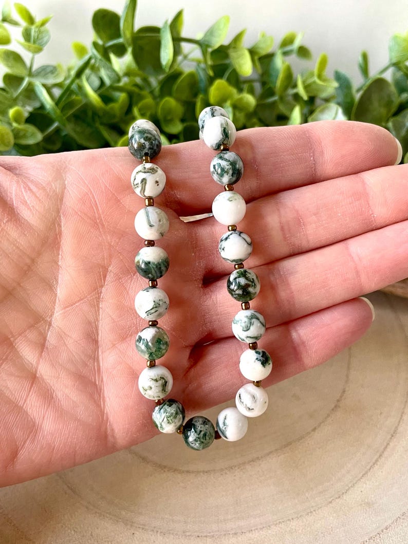 May include: A beaded bracelet featuring white and green moss agate stones. The round beads are interspersed with small gold-colored spacers. The bracelet is displayed against a background of green foliage.
