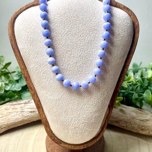 May include: A necklace of light blue, round beads with dark accents is displayed on a wooden jewelry bust. The beads have a marbled appearance. The bust is set against a neutral background with greenery.