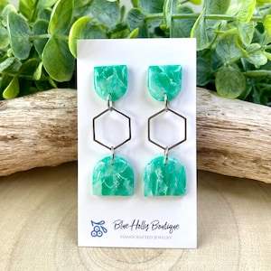 May include: A pair of handmade earrings with a marbled green design. The earrings feature a half-circle shape at the top and bottom, connected by a silver geometric shape. The earrings are displayed on a white card with the text "Blue Holly Boutique HANDCRAFTED JEWELRY".