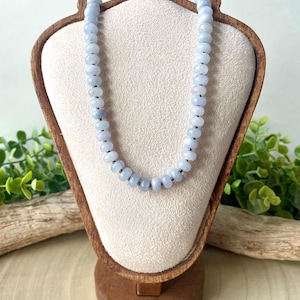 May include: A light blue beaded necklace displayed on a wooden jewelry stand. The necklace features round, light blue beads strung together with small, dark gray spacers. The stand has a light-colored fabric backing.