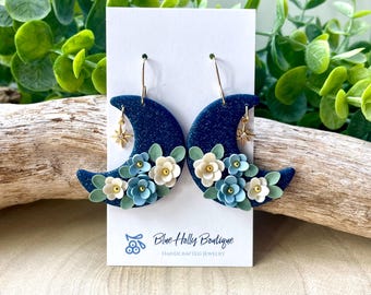 Op de afbeelding: Een paar donkerblauwe, glinsterende oorbellen in de vorm van een halve maan, versierd met kleine witte en lichtblauwe bloemen en een gouden sterbedel, gepresenteerd op een witte kaart met de tekst Blue Holly Boutique Handcrafted Jewelry.