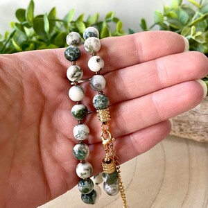 May include: A beaded bracelet featuring round, white and green gemstone beads. The bracelet has gold-colored metal accents and a decorative charm. The clasp is also gold-colored. The bracelet is held against a background of green foliage.