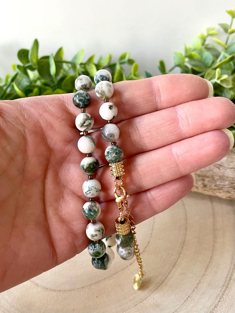 May include: A beaded bracelet featuring round, white and green gemstone beads. The bracelet has gold-colored metal accents and a decorative charm. The clasp is also gold-colored. The bracelet is held against a background of green foliage.