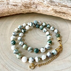 May include: A layered necklace featuring round, white and green patterned beads. The necklace has gold-colored accents and a small, round charm. The beads are strung on a delicate chain, creating an elegant accessory.