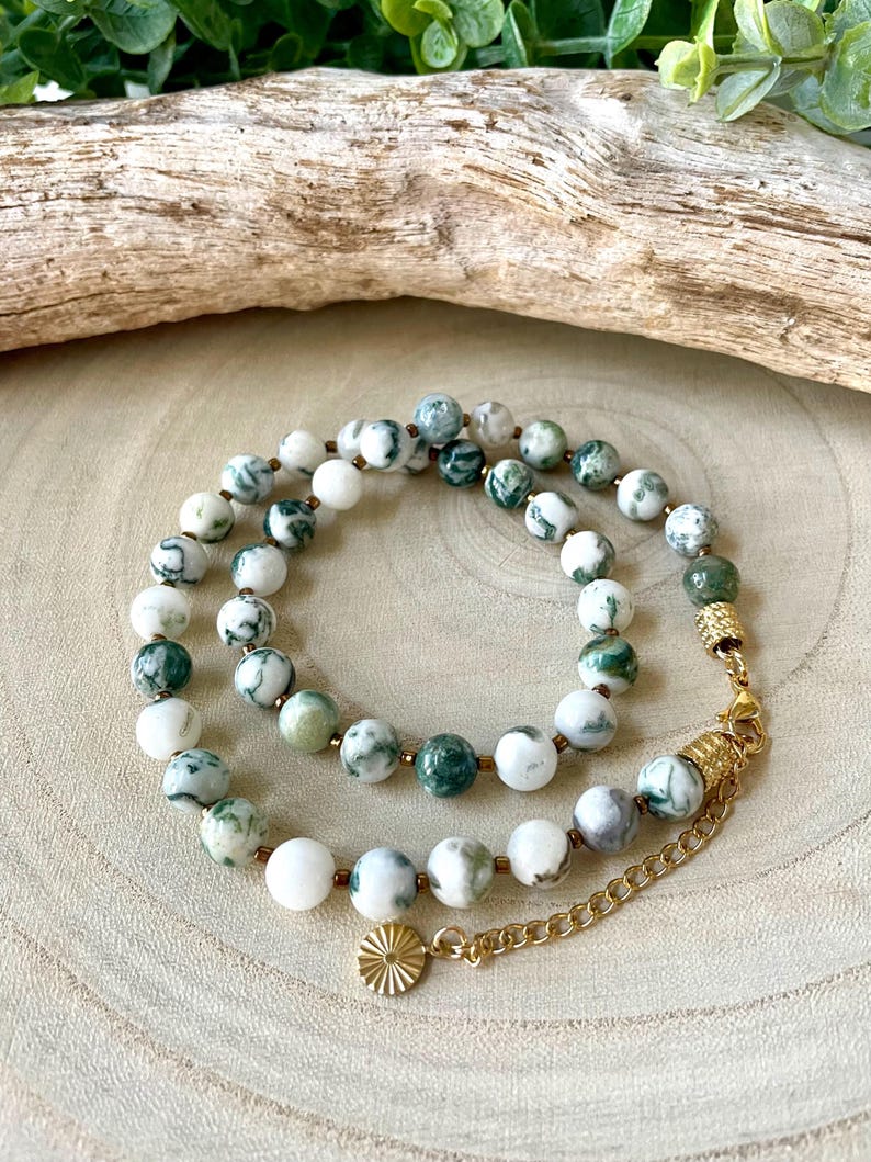 May include: A layered necklace featuring round, white and green patterned beads. The necklace has gold-colored accents and a small, round charm. The beads are strung on a delicate chain, creating an elegant accessory.