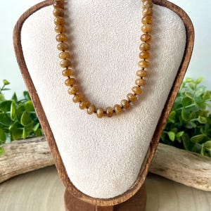 May include: A necklace with small, round, amber-colored beads. The beads are strung together, forming a necklace that is displayed on a wooden jewelry stand with a cream-colored fabric backing. The stand is set against a backdrop of greenery.