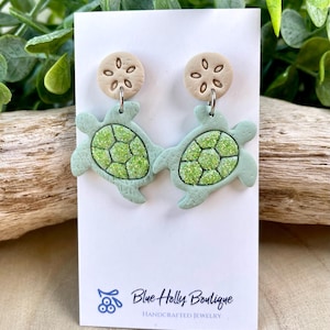 May include: A pair of handcrafted earrings featuring a sea turtle design. The earrings have light green turtle shapes with glittery green shells, and tan sand dollar studs. The earrings are displayed on a white card with the text "Blue Holly Boutique".