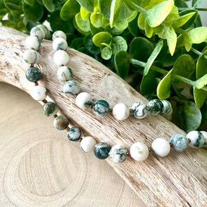May include: A beaded necklace featuring round, white and green patterned beads. The necklace is strung on a thin, gold-colored chain. The beads are interspersed with smaller, gold-colored beads. The necklace is displayed on a piece of driftwood.
