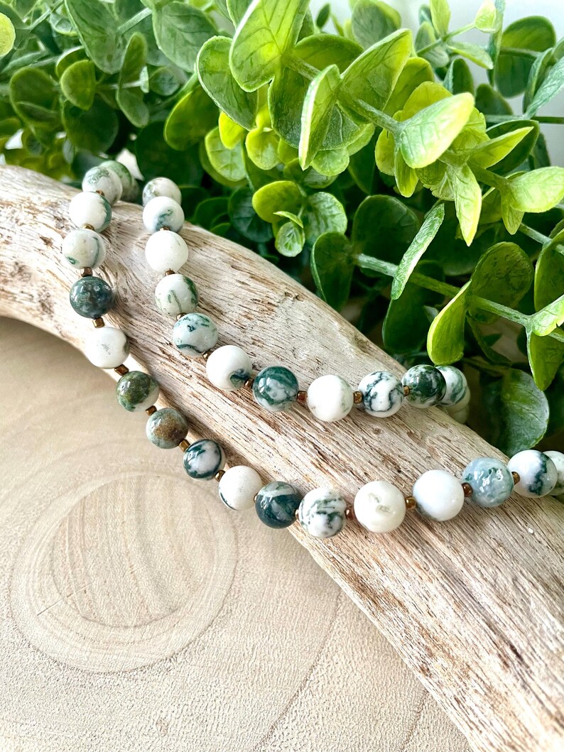 May include: A beaded necklace featuring round, white and green patterned beads. The necklace is strung on a thin, gold-colored chain. The beads are interspersed with smaller, gold-colored beads. The necklace is displayed on a piece of driftwood.