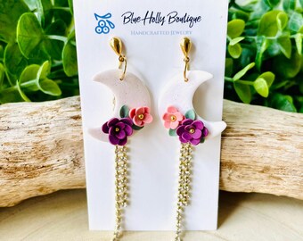 Op de afbeelding: Handgemaakte oorbellen met een halve maan ontwerp, versierd met roze en paarse bloemen en gouden kettingen. De oorbellen worden gepresenteerd op een witte kaart met de tekst "Blue Holly Boutique HANDCRAFTED JEWELRY".