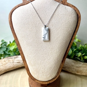 May include: A rectangular white and gray marble pendant necklace on a silver chain. The pendant is displayed on a cream-colored jewelry display stand with a wooden base. Green foliage and a piece of driftwood are in the background.