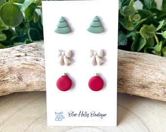 May include: A set of three pairs of Christmas-themed stud earrings on a white card. The earrings include green Christmas trees, cream-colored bows, and red ornaments with gold tops. The card has the text "Blue Holly Boutique".