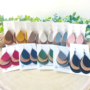 以下が含まれることがあります： 茶色、黄色、グレー、白、青、ピンク、赤、緑、黒など、さまざまな色のレイヤードティアドロップイヤリングのコレクション。 各ペアは革のトップレイヤーとコルクのボトムレイヤーを備え、白いカードに表示されています。