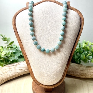 May include: A beaded necklace with light blue-green stones and gold-colored spacers. The necklace is displayed on a wooden jewelry stand with a cream-colored fabric backing. The stand is set against a neutral background with greenery.