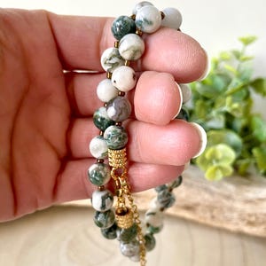 May include: A double-strand bracelet featuring round beads in shades of white, green, and gray. The beads are strung together with gold-colored accents and a delicate chain.