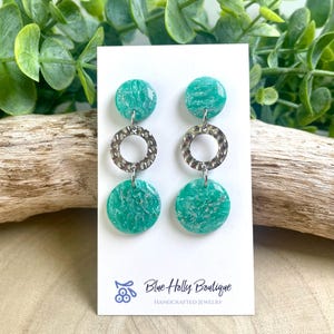 May include: A pair of handcrafted earrings with teal green circular pendants and textured silver rings. The earrings are displayed on a white card with the text "Blue Holly Boutique HANDCRAFTED JEWELRY". The earrings are set against a backdrop of greenery and driftwood.