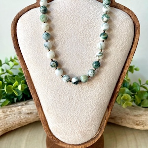 May include: A beaded necklace with green and white speckled stones. The necklace is displayed on a wooden jewelry stand with a beige fabric backing. The beads are round and appear to be natural stone. The necklace is a simple, elegant piece.