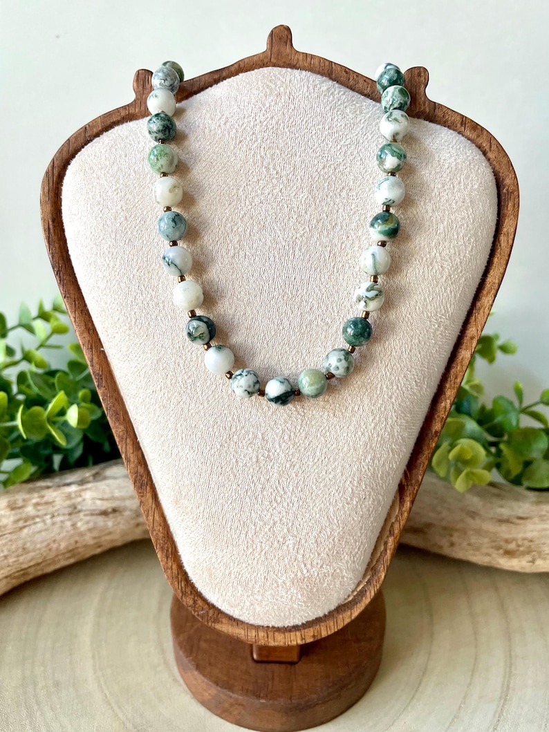 May include: A beaded necklace with green and white speckled stones. The necklace is displayed on a wooden jewelry stand with a beige fabric backing. The beads are round and appear to be natural stone. The necklace is a simple, elegant piece.