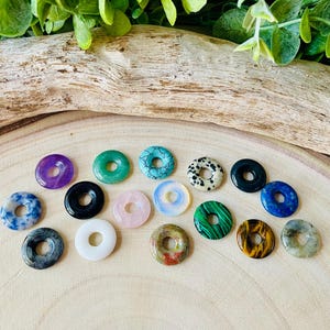 Könnte beinhalten: Eine Sammlung verschiedener Donut-Anhänger aus Edelsteinen in den Farben Lila, Grün, Blau, Schwarz, Weiß, Rosa und Gelb. Die Anhänger sind auf einer hellen Holzoberfläche angeordnet und zeigen ihre einzigartigen Farben und Muster.