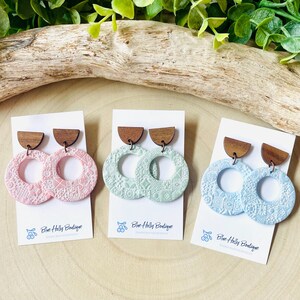 May include: Three pairs of handmade earrings in pastel colors: pink, mint green, and light blue. Each pair features a wooden half-circle top and a textured, circular pendant with a detailed pattern. The earrings are displayed on white cards.