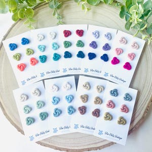 May include: A collection of colorful knot-shaped stud earrings displayed on white cards. The earrings come in various colors, including blue, green, pink, and neutral tones. The cards are arranged on a wooden slice, with greenery in the background. The text "Blue Holly Boutique" is visible.