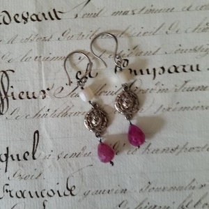 Op de afbeelding: Een paar zilveren oorbellen met roze en witte stenen. De oorbellen hebben een bloemmotief en hangen aan een zilveren haak.