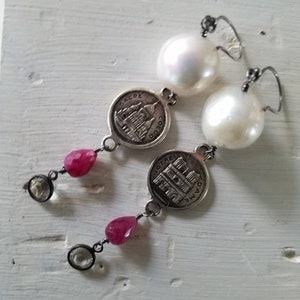 May include: A pair of silver earrings with a pearl and a charm featuring the Sacre Coeur Basilica in Paris. The charms are attached to a red gemstone and a clear gemstone.