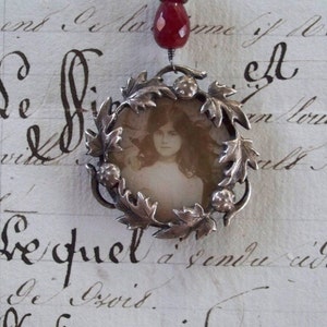Peut inclure: Un collier en argent avec un pendentif en pierre rouge et un médaillon en argent contenant une photo d'une jeune fille. Le médaillon est entouré d'un motif de feuilles en argent.