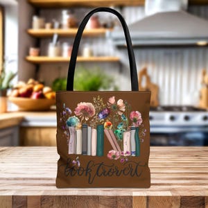 Op de afbeelding: Bruine tote bag met een bloemenpatroon en een stapel kleurrijke boeken. De tekst "booktrovert" is op de tas gedrukt.