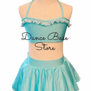 May include: A light blue two-piece dance outfit on a mannequin, featuring a halter-style crop top with a ruffled neckline and a matching pleated skirt. The text "Dance Base Store" is printed in black script across the center of the top.