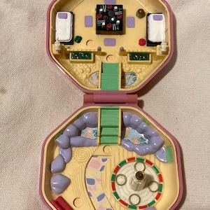 Puede incluir: Una casa de juguete de plástico rosa con una parte superior extraíble. La parte superior revela una sala de estar en miniatura con una cama, una mesa y una chimenea. La parte inferior de la casa muestra un área exterior en miniatura con una fuente, un banco y un camino.