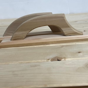 May include: Two wooden hand planes with arched handles and rectangular bases. The planes are made of light-colored wood and are positioned on a light-colored wooden surface. The handles are curved, providing a comfortable grip for woodworking tasks.