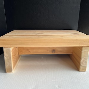 May include: A small, rectangular wooden step stool. The stool is made of light-colored wood and has a simple, sturdy design. The top is made of horizontal planks, and the legs are solid blocks. The stool measures approximately 12 inches long, 10 inches wide, and 6 inches high.