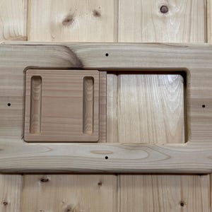 May include: A rectangular wooden frame with a recessed area and a sliding panel. The frame is made of light-colored wood and has rounded corners. The sliding panel has two vertical grooves. The wood grain is visible.