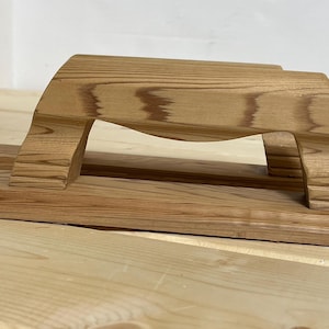 May include: A wooden ironing board press with a handle. The press is made of light brown wood with visible grain patterns. The handle is arched, and the base is rectangular. The press is designed for pressing seams and hems.