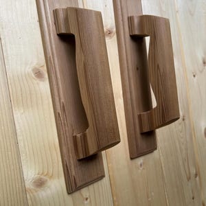 May include: Two wooden door handles with a rectangular backplate and a curved handle. The handles are made of light brown wood and are mounted on a light-colored wooden surface. The handles are designed for easy gripping.